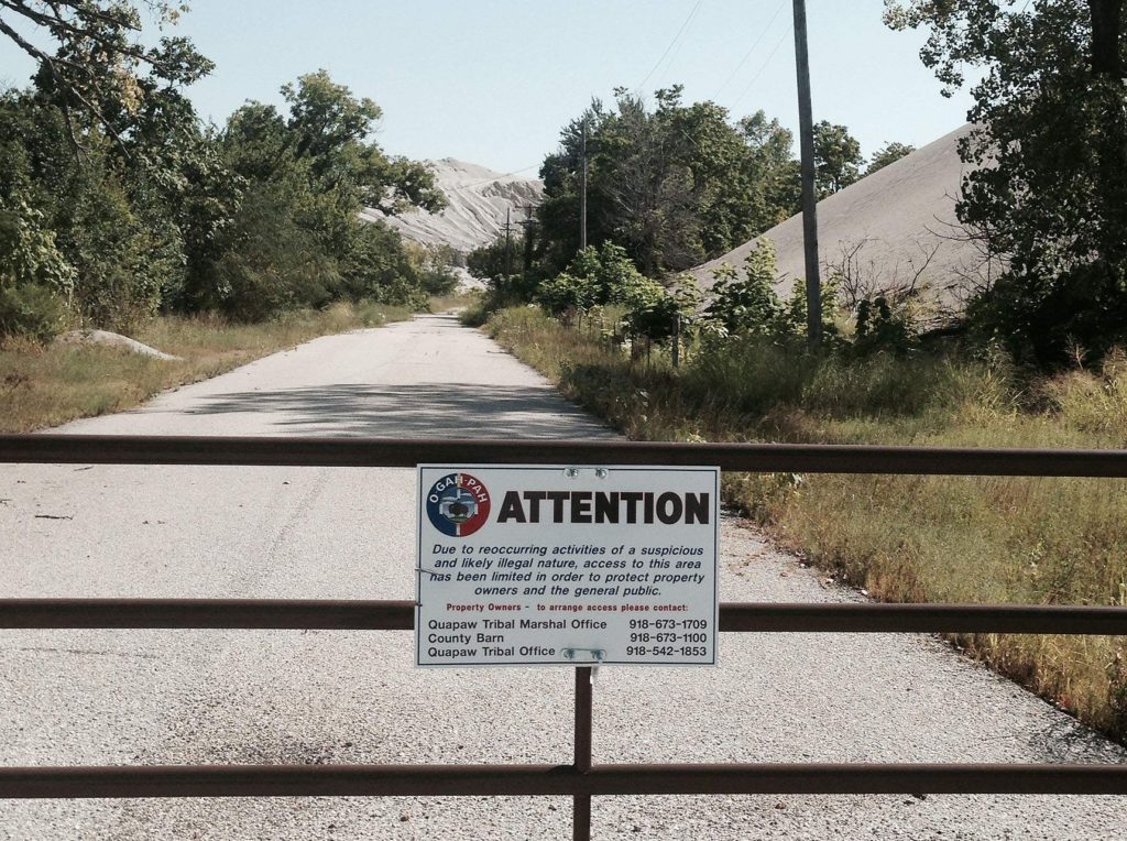 A town road in Picher, Okla. is blocked by the Quapaw Tribe on Aug. 23, 2014. A few of the massive chat piles that surround the community can be seen in the distance. Other roads are blocked by the federal government, and both entities are working together to begin clean up of the Tar Creek Superfund site. In Dec. 2013, the U.S. Environmental Protection Agency entered into its first ever agreement with a tribe, awarding the Quapaw $2.6 million to help with the clean up of a 40-acre area polluted by mining southeast of the town of Quapaw. Much of the Superfund site is owned by the Quapaw, a portion of the reservation they were given when they were forced from their original lands. (Lynda Waddington/The Gazette)